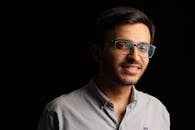 Close-up portrait of a young man wearing eyeglasses, smiling against a black background.